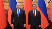 Mijaíl Mishustin y Li Qiang de pie, uno junto al otro, ante banderas de Rusia y China