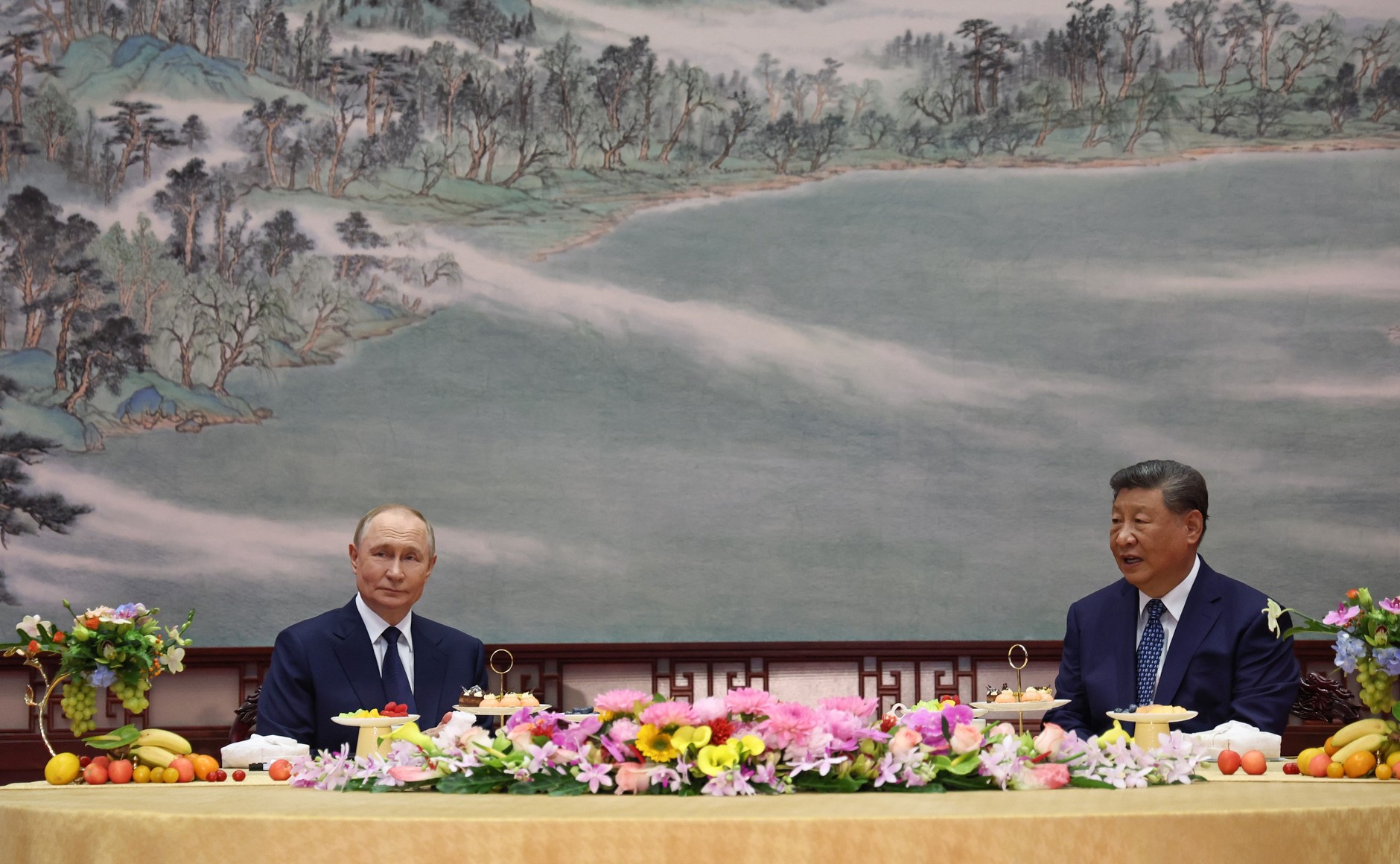 Vladímir Putin y Xi Jinping sentados a una mesa con flores ante un mural de paisaje en la residencia de Zhongnanhai, Pekín